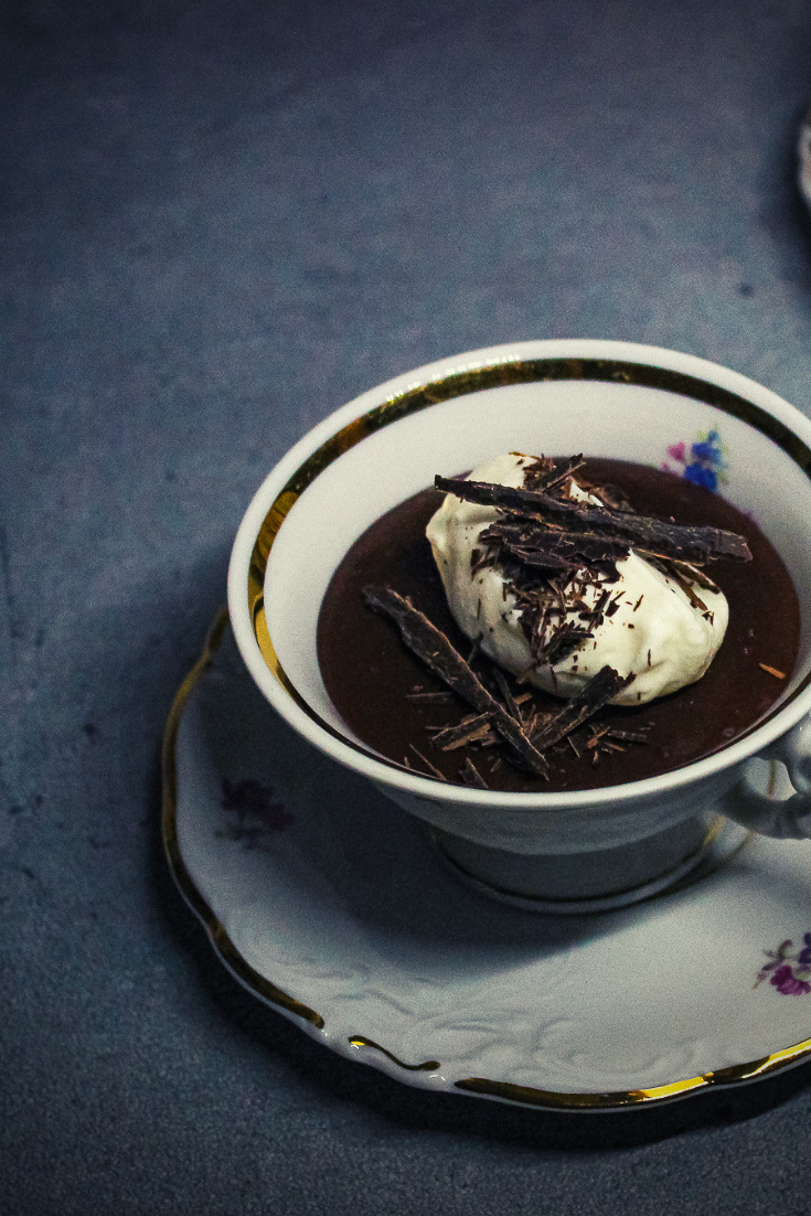 Dieses Bild zeigt eine Porzellantasse mit Goldrand und Blüten gefüllt mit einem frisch gekochten Schokoladenpudding. Dekoriert ist dieser mit Schlagsahne und Schokoladensplittern.